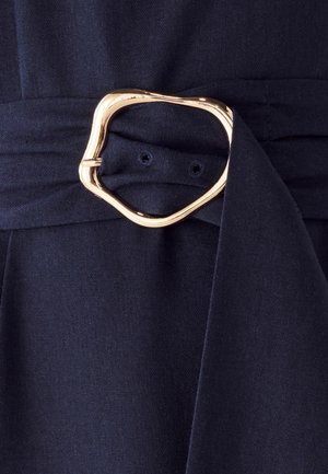 Navyblauwe stof met een gladde textuur, voorzien van een grote, abstracte gouden metalen gespsluiting op een strikdetail rond de taille.
