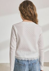Maglietta bianca a maniche lunghe con un motivo a diamante testurizzato e bordo in pizzo all'orlo, abbinata a pantaloni corti di jeans blu, vista da dietro.