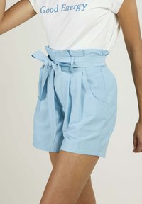 Shorts taille haute bleu clair avec une taille à nouer, un devant plissé et des poches latérales, fabriqués dans un tissu doux adapté à un style décontracté.