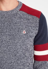 Pull tricoté avec un corps bleu marine chiné, épaules texturées rouge et bleu foncé, accents rayés blancs et rouges sur les manches, petit détail de logo.