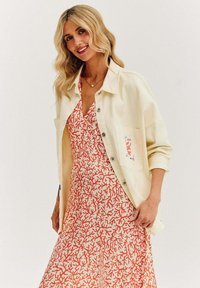 Krémová button-up bunda cez červené a biele šaty s koralovým vzorom. Šaty majú výstrih do V. Bunda má detail vrecka.