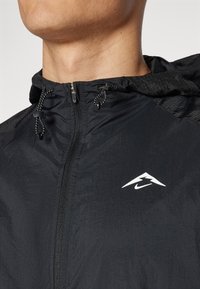 Czarna lekka kurtka z zamkiem z przodu, regulowanym kapturem ze sznurkiem i białym logo Nike na klatce piersiowej. Teksturowany detal materiału na ramionach.