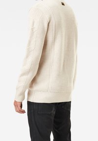 Cream-colored sweater with ribbed texture, featuring cable-knit patterns on sleeves and a small logo at the neckline. Black pants are worn.