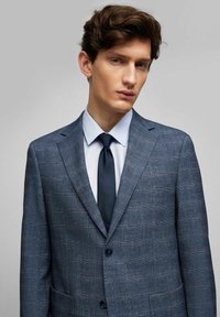 Navyblauwe geruite blazer met twee knopen, gecombineerd met een lichtblauwe blouse en een donkere stropdas, die een soepele textuur en een getailleerde pasvorm laat zien.
