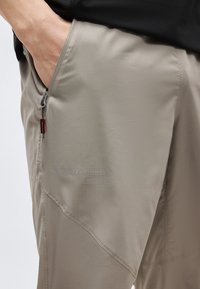 Hånd i lomme på beige utendørsbukse med glidelåslomme og subtil merkevarelogo på lårpartiet.