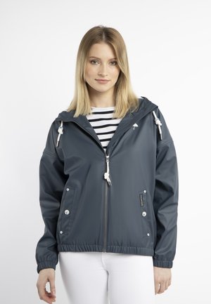 Jeune femme portant une veste à capuche zippée gris foncé sur un t-shirt rayé noir et blanc et un pantalon blanc devant un fond uni.
