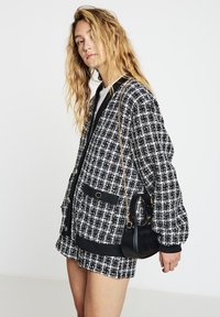 Schwarze-weiße Bomberjacke mit kariertem Muster, lockerem Schnitt, gerippten Abschlüssen und zwei Vordertaschen, kombiniert mit einer kleinen schwarzen Handtasche.