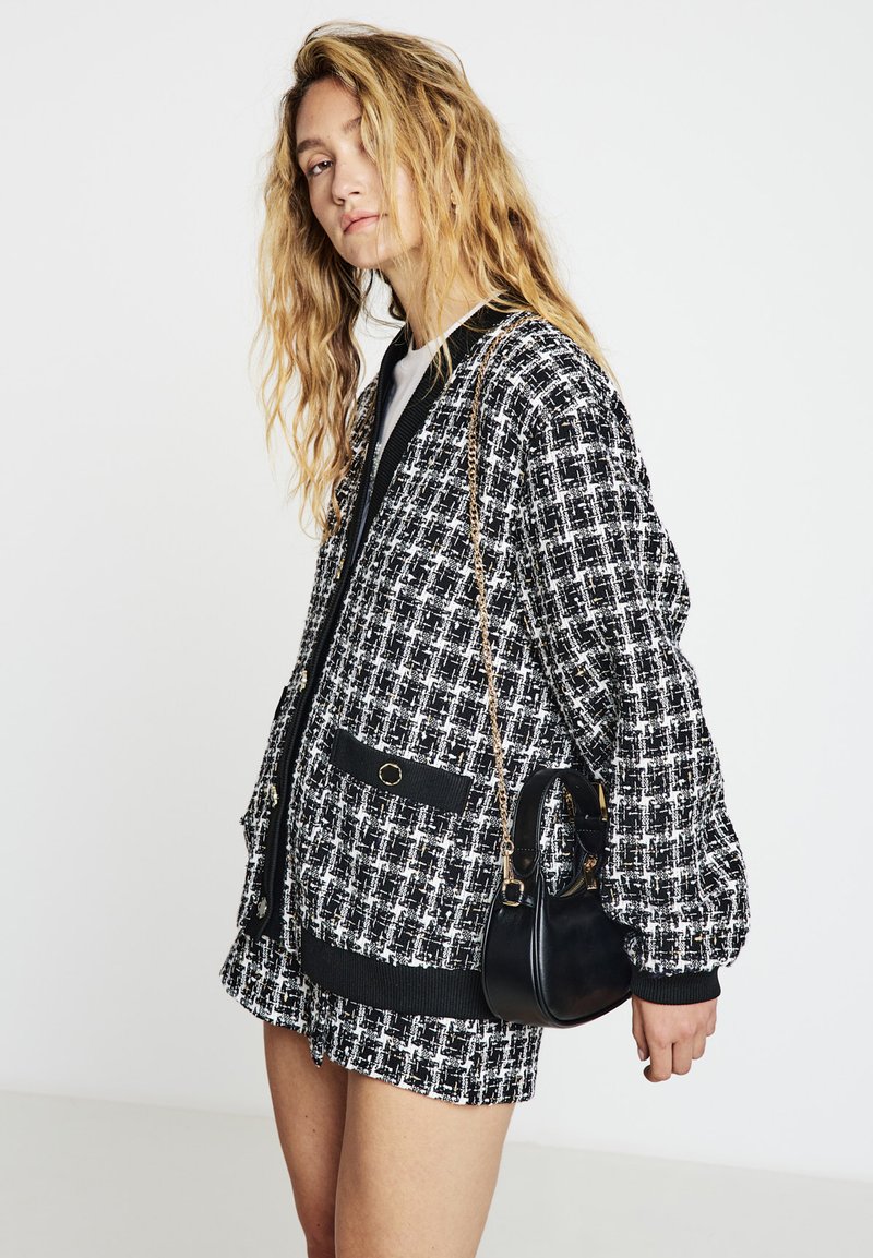 Schwarze-weiße Bomberjacke mit kariertem Muster, lockerem Schnitt, gerippten Abschlüssen und zwei Vordertaschen, kombiniert mit einer kleinen schwarzen Handtasche.