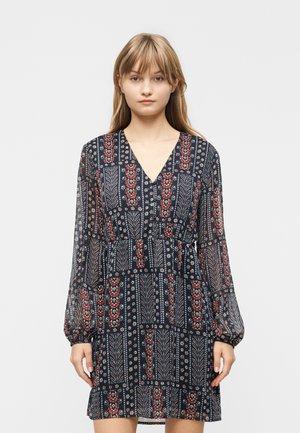 Joven con un vestido de manga larga, estampado, hasta la rodilla, con escote en V y cintura entallada, posando frente a un fondo liso.