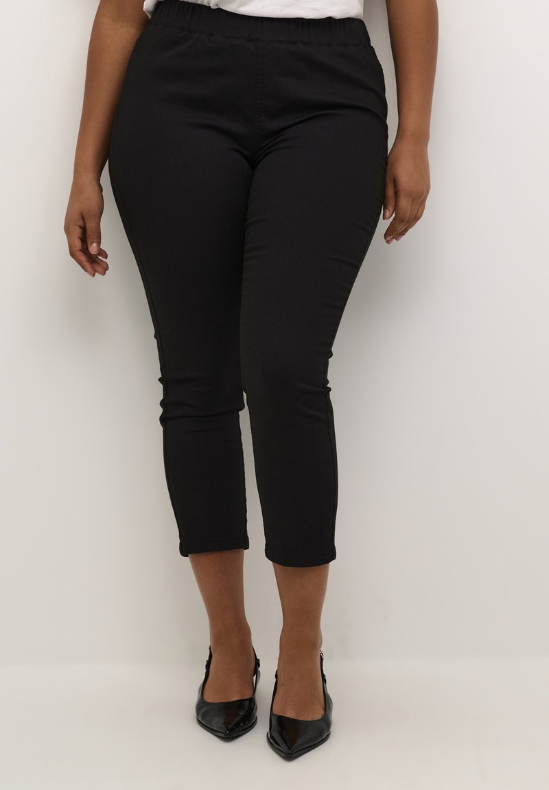 Pantaloni neri lungo caviglia con fascia elastica, texture liscia e design aderente, abbinati a tacchi a punta neri.