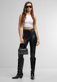 Pantalon noir taille haute en similicuir avec fermetures éclair, associé à un débardeur court blanc et un sac à main noir ; modèle portant des lunettes de soleil noires oversize.