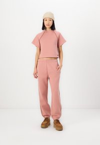 Rosa kurz geschnittenes Sweatshirt mit Raglanärmeln, kombiniert mit passenden Jogginghosen mit elastischen Bündchen und seitlichen Taschen. Das Modell trägt eine beige Mütze.