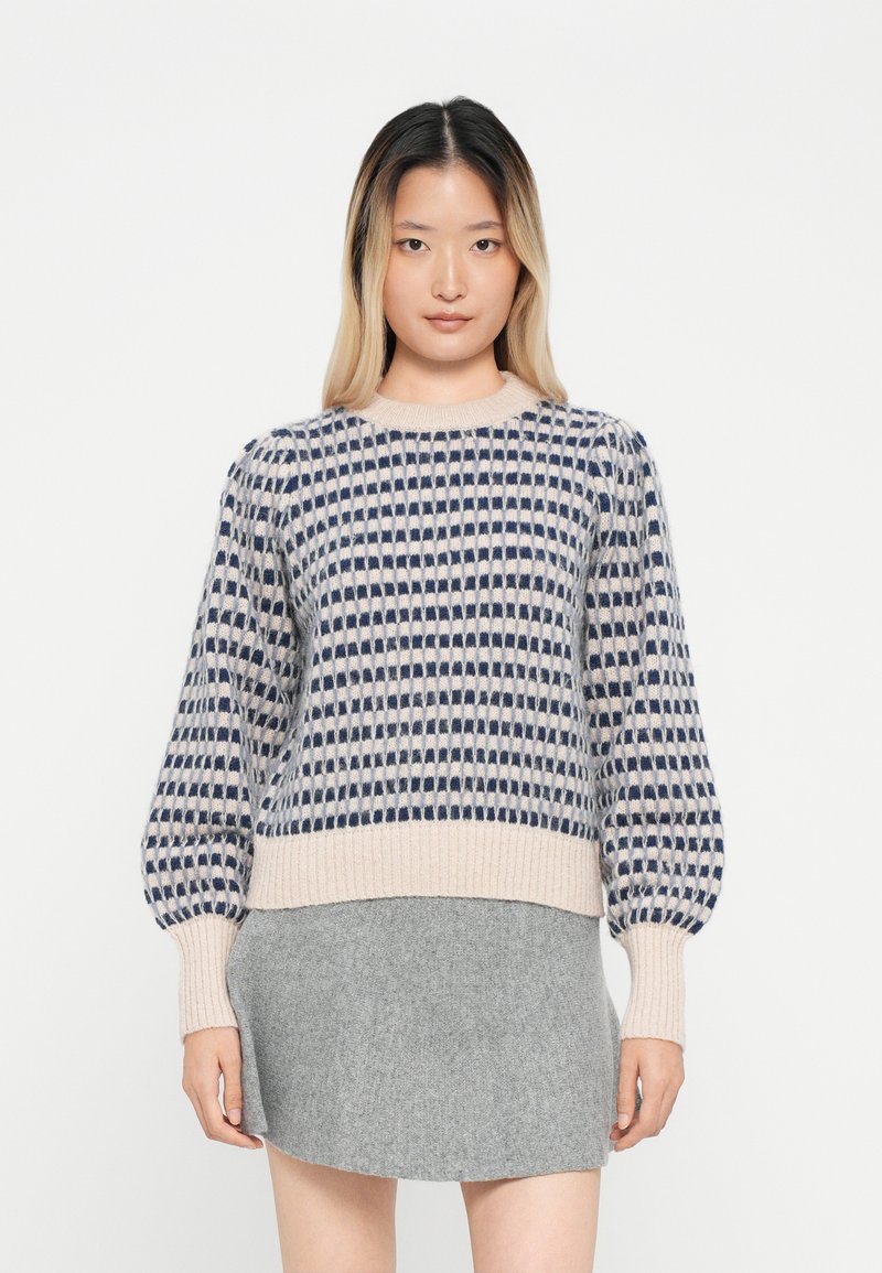 Strickpullover mit einem cremefarbenen und marineblauen Karomuster, gerippten Bündchen und einem runden Ausschnitt. Kombiniert mit einem grauen Rock.