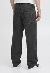Jeans de mezclilla negra con un corte suelto, textura desgastada, dos bolsillos traseros y un diseño de pierna recta, combinados con zapatillas blancas.