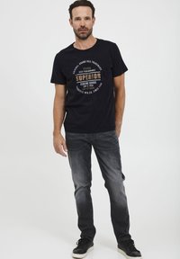 Melns kokvilnas T-krekls ar apaļu grafisko druku bēšā un baltā krāsā, apvienots ar gaiši pelēkiem džinsiem un melnām sporta apaviem.