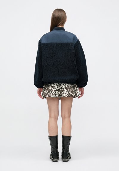 Γυναίκα που στέκεται ανάποδα φορώντας ένα ναυτικό fleece μπουφάν, κοντή φούστα με εκτύπωση λεοπάρδαλη και μαύρες μπότες μέχρι τη μέση της γάμπας με λεπτομέρειες αλυσίδας.