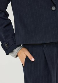 Námořnický blazer s proužky vyrobený z texturované látky s klopovým límcem, zapínáním na knoflíky a viditelnými šedými rukávy s manžetou.