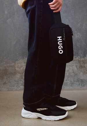 Černé kožené tenisky s bílou podešví, černé džíny s kontrastním prošitím a černá taška s bílým logem "HUGO".