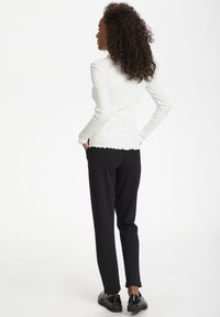 Haut long à manches ribbées blanches avec ourlet festonné associé à un pantalon noir et des chaussures noires. Le modèle est vu de dos.