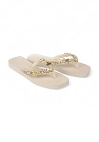 Beige teenslippers met een gestructureerde zool, voorzien van een decoratieve band versierd met gouden accenten, parels en schelpenontwerpen.