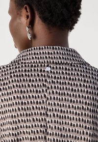 Blouse with a grey and beige pattern featuring black dots, fabric texture is smooth, includes a collar and a small back button detail.
