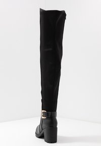 Bottes synthétiques noires montant jusqu'au genou, à finition lisse, forme effilée, talon bloc et accent de boucle à la cheville. Détail de fermeture éclair sur le côté.