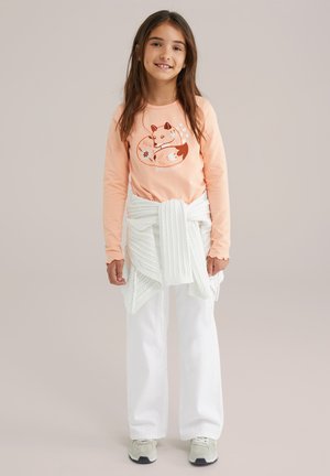 Licht perzikkleurig langemouwen T-shirt met een vosafbeelding, gecombineerd met witte wijde broek en een gebreide witte trui die om de taille is geknoopt.