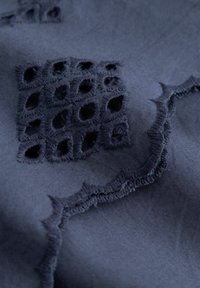 Ciemnoniebieska tkanina z misternie haftowanymi wycięciami tworzącymi wzór z małych, diamentowych otworów i zakrzywionych linii.