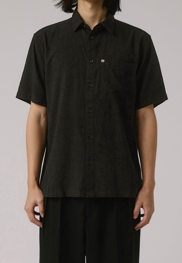 Homme portant une chemise noire à manches courtes boutonnée avec un motif subtilement texturé et une poche poitrine, associée à un pantalon noir ample.