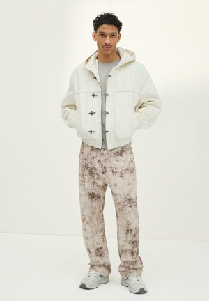 Młody mężczyzna stojący z rękami w kieszeniach, ubrany w kremową kurtkę z kapturem, szarą koszulę, wzorzyste luźne spodnie i białe trampki.
