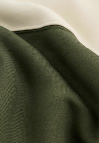 Kampsuni kangas, mis on oliivrohelise ja kreemja värviga, sujuva tekstuuriga ning selgete õmblusjoontega värviblokkide vahel.