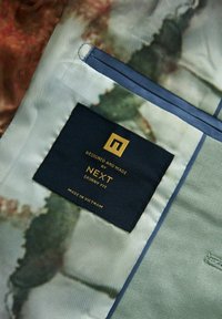Étiquette en tissu d'un vêtement avec un fond bleu foncé et un texte doré. Le matériau du vêtement présente un motif de couleur pastel.
