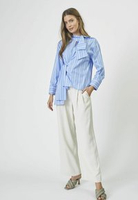 Hellbluse mit Streifen und Schleife, kombiniert mit weißen weiten Hosen und grünen Absatzsandalen, vor einem schlichten Hintergrund.
