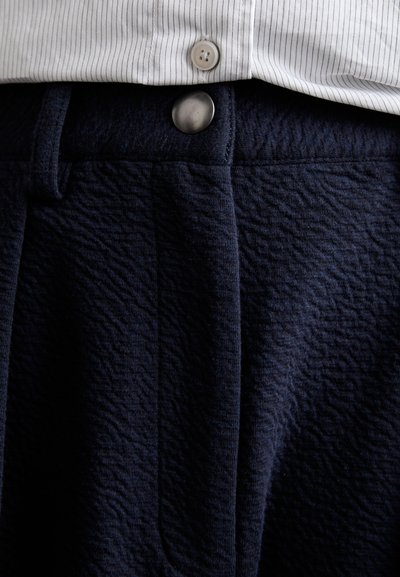 Pantaloni strutturati blu navy con un bottone metallico visibile in vita, abbinati a una camicia bianca a righe parzialmente visibile sopra.