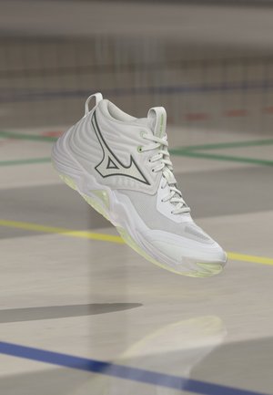 Witte sportschoen met zwart Mizuno-logo die boven een indoor sportveld zweeft met meerkleurige lijnen en een vage achtergrond.
