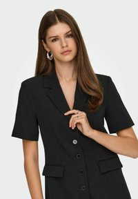 Crna krojena haljina s kratkim rukavima, v-izrezom, zatvaranjem na pet dugmadi i prednjim džepovima. Glatka tkanina s čistim linijama. Srebrne okrugle naušnice.