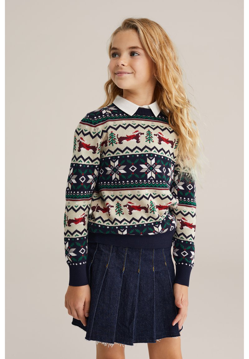 Stickad tröja med ett festligt mönster med röda rävar, gröna träd och snöflingor på en krämfärgad och marinblå bakgrund; kombinerad med en jeanskjol.