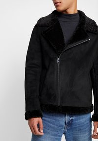 Mies, joka pukeutuu mustaan shearling-takkiin, jossa on pörröiset kaulus ja hihansuut, musta vetoketju, tumma poolopaita ja siniset farkut, seisoo yksinkertaista taustaa vasten.