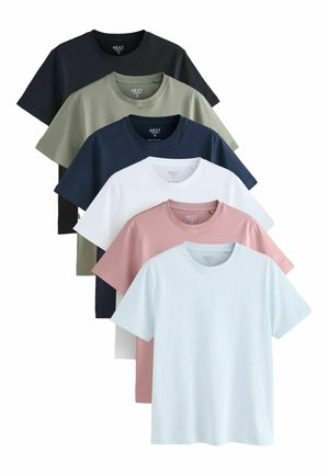 Homme aux cheveux courts et bouclés portant un t-shirt bleu clair et un jean bleu, debout devant un fond beige uni.