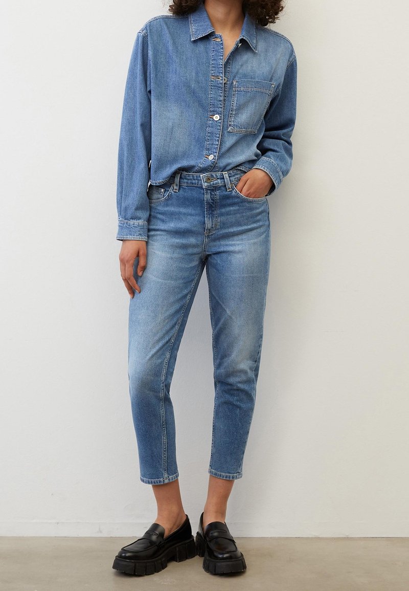 Personne portant une chemise en jean bleue et un jean bleu délavé taille haute, avec des mocassins à plateforme épaisse noirs, debout contre un mur blanc.
