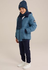 Blå puffjacka med marinblå detaljer, matchad med en mörkblå stickad halsduk och mössa. Modellen bär marinblå byxor och vita sneakers.