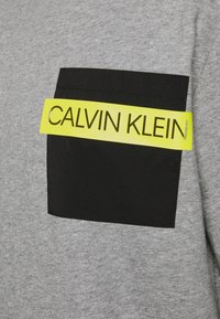 Tissu gris avec une poche rectangulaire noire surmontée d'une bande jaune affichant le texte "CALVIN KLEIN" en lettres majuscules.
