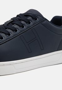 Námořnické kožené tenisky s kulatou špičkou, plochou bílou podrážkou a šněrovacím designem. Na boku se nachází perforované písmeno "H". Texturovaný povrch.