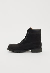 Must mustaankle saapa musta nahast, ümarate varvastega, pehmendatud kraega, metallidega silmadega ja tekstuuriga kummitaldadega. Küljel on nähtav õmbluse detail.