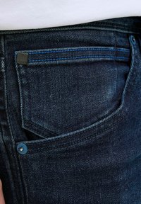 Jeans din denim de culoare albastru închis, cu un buzunar frontal vizibil și cusături în contrast. Prezintă o etichetă metalică și o cusătură albastră de accent pe buzunar.