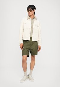 Ανοιχτό μπεζ τζάκετ από τζιν με κουμπιά, ελαιώδεις cargo σορτς, κρέμα sneakers και γυαλιά ηλίου. Απλός, λειτουργικός σχεδιασμός.