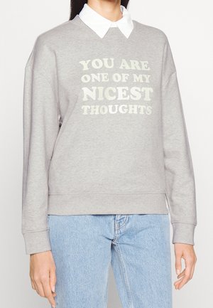 Person iført en grå sweatshirt med hvid tekst "DU ER EN AF MINE DEJLIGSTE TANKER" lagdelt over en hvid skjorte med krave og blå jeans.