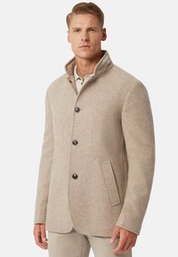 Abrigo de mezcla de lana beige con cuello alto, cinco botones negros, un bolsillo y un corte entallado. Textura suave y diseño estructurado.