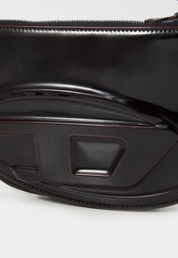 Zwarte patenten leren tas met een gladde, glanzende oppervlakte. Kenmerkend is het gebogen ontwerp, de ritssluiting en de rechthoekige hardwareaccenten.