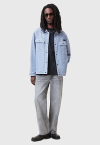 Chaqueta de pana azul claro con dos bolsillos en el pecho, camiseta negra debajo, vaqueros de pierna ancha gris, zapatos negros y gafas de sol.